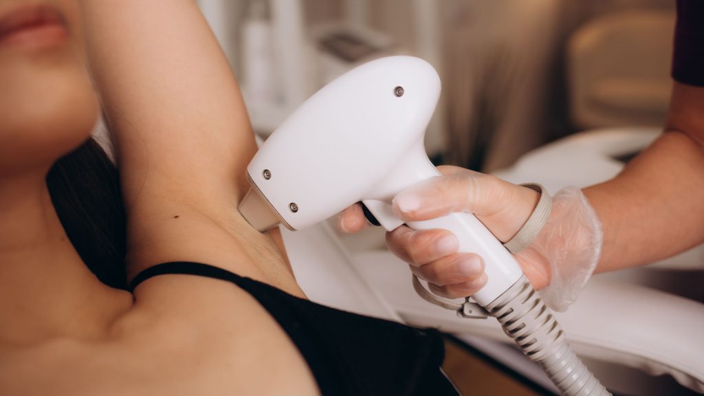 Body care concept. Laser hair removal. Young Caucasian woman in safety glasses lies on the sofa, female cosmetologist doing laser hair removal in a beauty salon